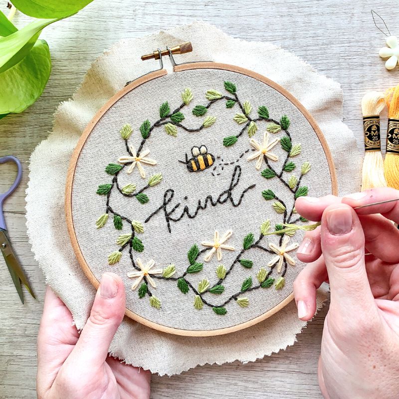 Person stitching a bee embroidery design next to heart-shaped sewing scissors, embroidery floss, and a green plant