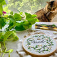 A bee embroidery hoop next to a plant and cat