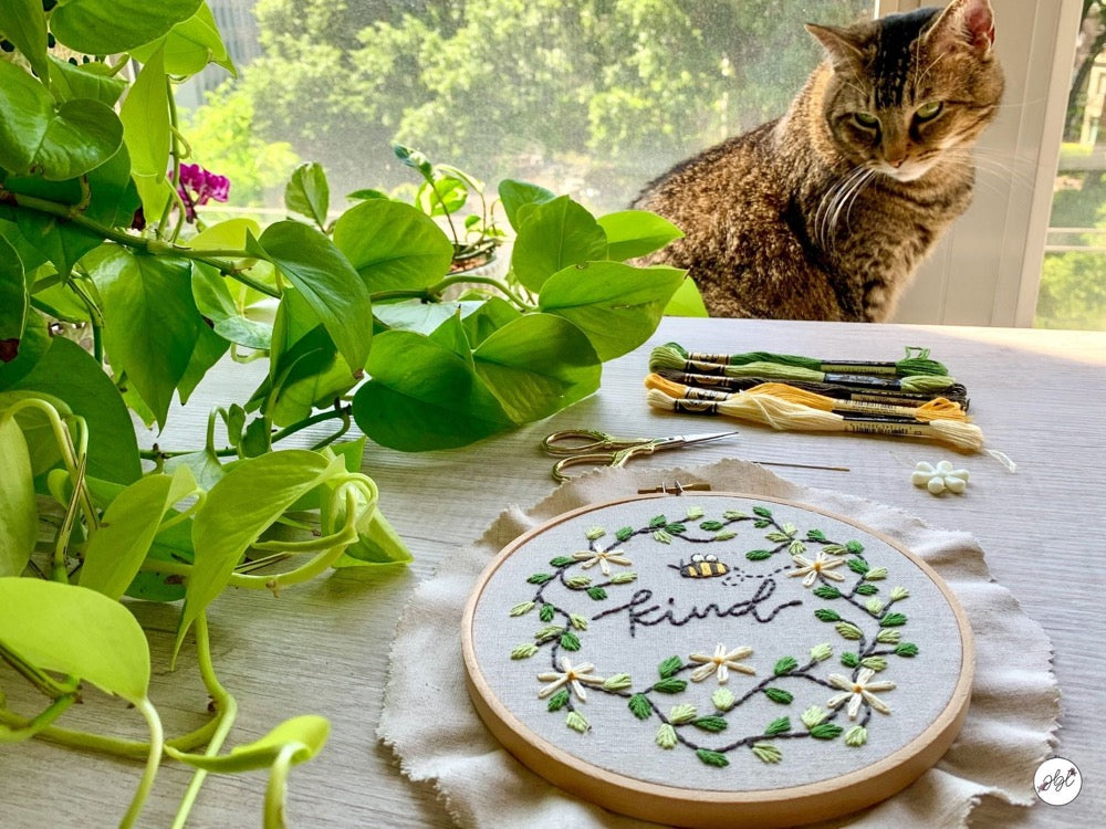 A bee embroidery hoop next to a plant and cat