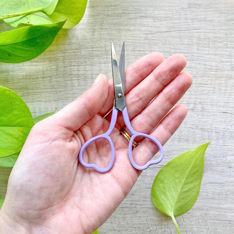 A hand holding purple embroidery scissors next to a green plant