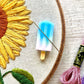 A blue and pink popsicle needle minder holding a needle on an embroidery hoop with an embroidered sunflower