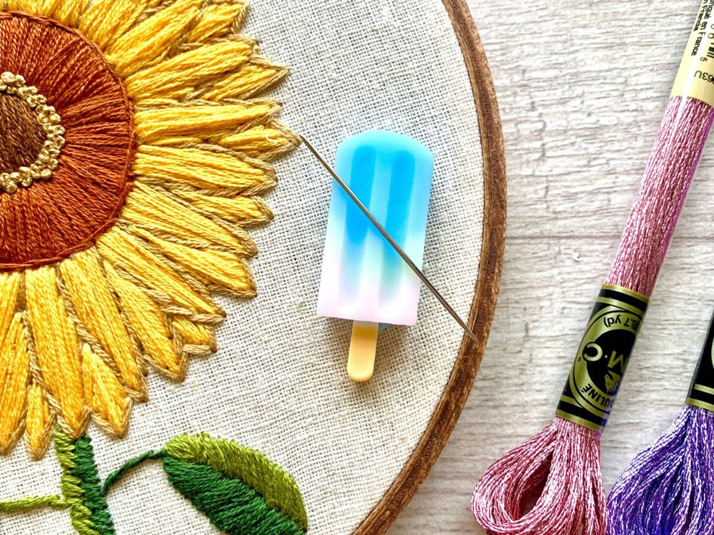A blue and pink popsicle needle minder holding a needle on an embroidery hoop with an embroidered sunflower